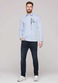 Helles blaues Hemd mit Knopfverschluss und gestreiftem Muster sowie schwarzem Textlogo, kombiniert mit dunklen Jeans und weißen Turnschuhen.