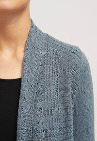 Stickad cardigan i mjuk blå färg, med ett texturerat mönster och öppen framsidan. Tyget verkar lätt och har en delikat, luftig design.