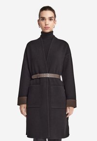 Schwarzer langer Mantel mit tailliertem Gürtel, zwei Eingrifftaschen und braun getönten Manschetten. Hergestellt aus einem weichen, wollähnlichen Material.