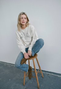 Krämfärgad sweatshirt med grafiskt tryck, blå jeans och bruna ankelboots, sittande på en träpall mot en neutral vägg.