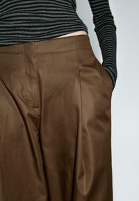 Osoba v hnědých kalhotách s vysokým pasem, s rukama v kapsách a v černém a šedém pruhovaném topu s dlouhým rukávem na jednobarevném pozadí.