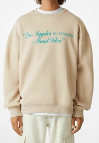 Sweatshirt beige oversize con polsini e orlo a coste. Presenta testo turchese: "Los Angeles È SEMPRE UNA Buona Idea". Tessuto morbido.