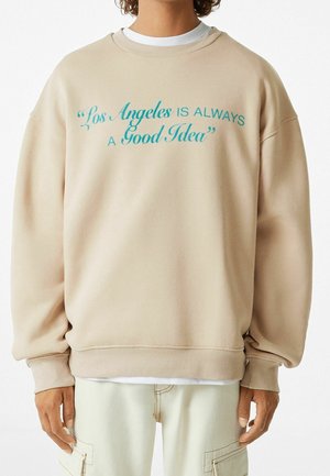 Sweatshirt beige surdimensionné avec des poignets et un ourlet côtelés. Présente du texte bleu canard : « Los Angeles EST TOUJOURS Une Bonne Idée. » Texture de tissu douce.