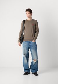 Kaki genser med lange ermer og rødt logo på brystet, kombinert med lyseblå ripped jeans og svarte sko.