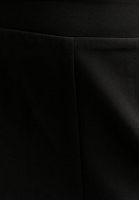 Tela negra con una textura suave y mate. Presenta costuras horizontales y un leve brillo. Cortada en un diseño entallado y estructurado.