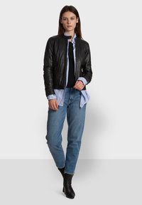 Crna kožna bomber jakna s uzdignutim ovratnikom, uparena s crnim topom i plavom prugastom košuljom, nosi se preko svijetloplavih traperica i crnih gležnjača.