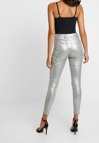 Mujer con pantalones ajustados plateados brillantes, top negro con tirantes y sandalias negras de tacón alto, de pie contra un fondo blanco liso.