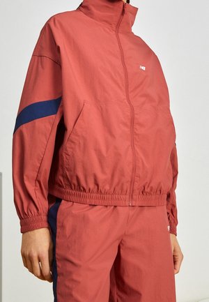 Person iført en koralrød træningsdragt med marineblå striber på ærmer og bukser, stående mod en lysegrå baggrund.