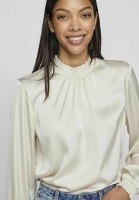 Creme satinbluse med høj flæsekrave, lange ærmer og samlede manchetter, båret med denimjeans; glat tekstur og elegant design.