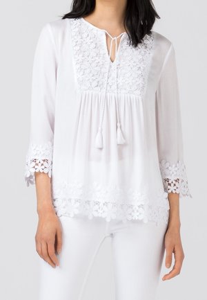 Witte blouse met lange mouwen en bloemrijke kanten inzetstukken op de borst, manchetten en zoom, voorzien van een halslijn met kwastjes en een losse pasvorm op een persoon.