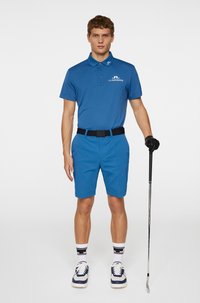 Blå pikétröja med logotyp, matchande blå shorts, svart bälte, vit och blå sneakers, vita strumpor med svarta accenter, hållandes en golfklubba.