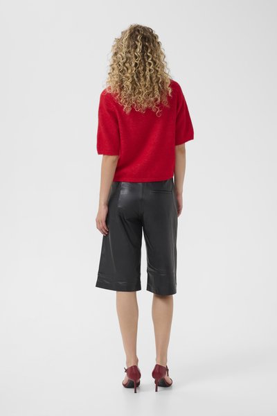 Femme aux cheveux blonds bouclés portant un haut rouge à manches courtes, un short en cuir noir jusqu'aux genoux et des chaussures à talons hauts rouges, vue de dos.