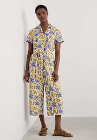 Blommönstrad jumpsuit i gult med blåa och gröna mönster, korta ärmar och midjebälte; vida ben och lätt material.