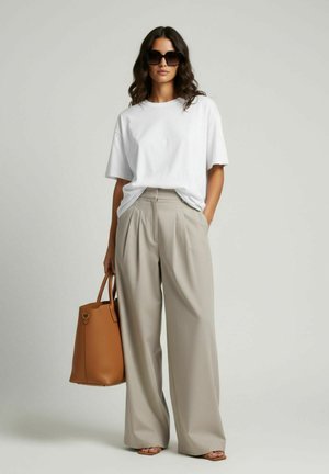 Vrouw draagt een oversized wit T-shirt, beige wijde broek, grote zonnebril en een bruine handtas, staand tegen een effen achtergrond.