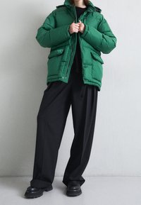 Veste doudoune verte avec capuche, présentant un design matelassé, des poches avant et des boutons-pression. Associée à un pantalon noir ample et des bottes noires épaisses.