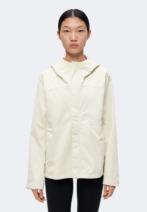 Jeune femme aux cheveux foncés attachés, portant une veste à capuche beige clair et un pantalon noir, face à un fond uni.