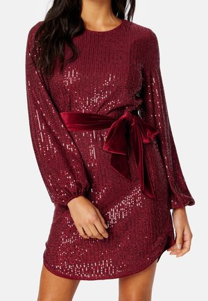 Femme portant une robe bordeaux à sequins à manches longues avec une ceinture en velours nouée à la taille.