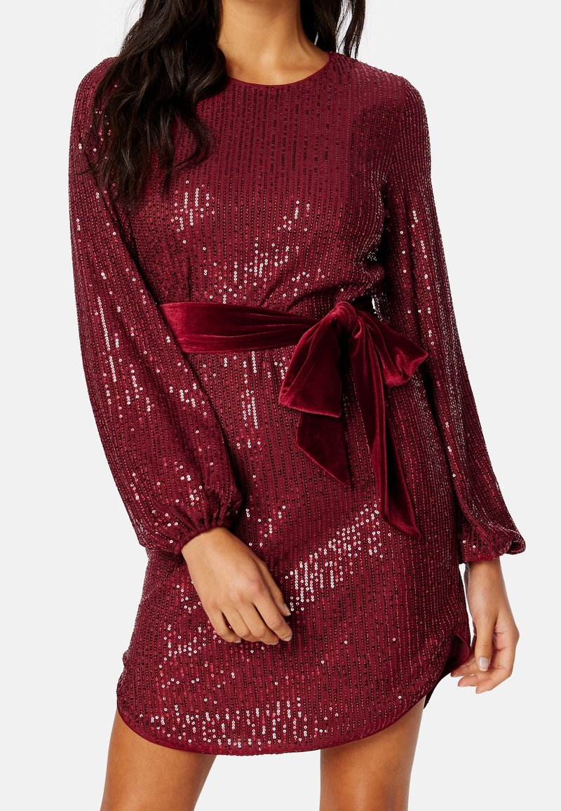 Femme portant une robe bordeaux à sequins à manches longues avec une ceinture en velours nouée à la taille.