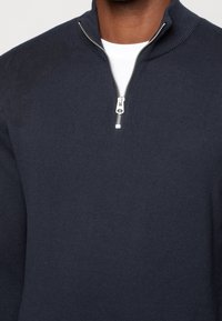 Pullover navy con colletto alto e chiusura a mezza zip in argento. Tessuto lavorato a maglia, texture liscia, design minimalista.