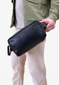 Zwarte leren toilettas met een rechthoekige vorm, ritssluiting en een kort handvat; gladde textuur met een reliëflogo-detail.