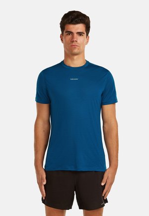 Uomo in piedi, volto rivolto in avanti, che indossa una maglietta atletica blu a maniche corte e pantaloncini neri su uno sfondo bianco uniforme.