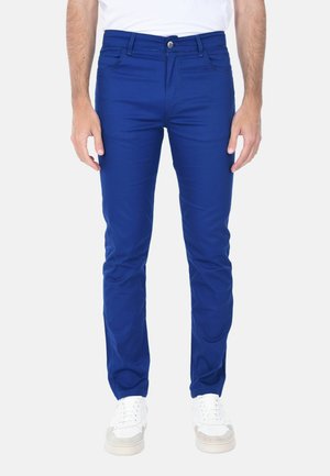 Uomo con pantaloni slim fit blu reale e scarpe da ginnastica bianche, in piedi con le braccia rilassate lungo i fianchi davanti a uno sfondo semplice.