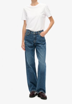 Person iført en enkel hvid kortærmet t-shirt, stoppet ned i højtaljede, vide blå jeans med brune sko. Den ene hånd i lommen.