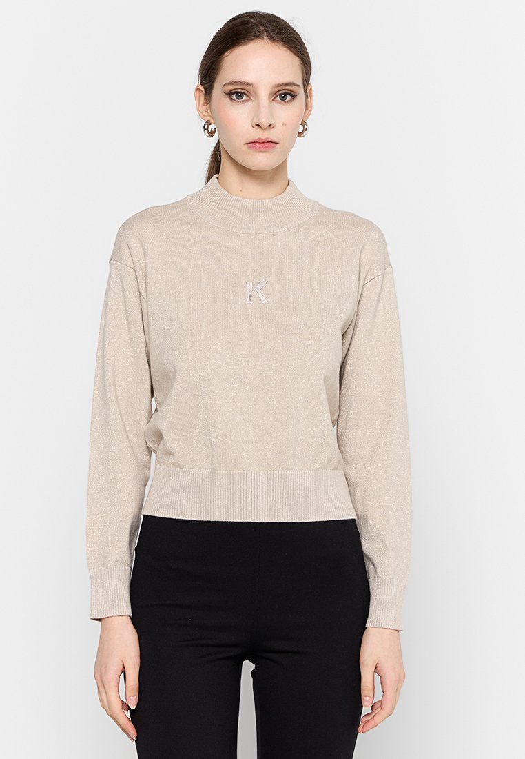 Donna che indossa un maglione beige a maniche lunghe con il logo "K" sul petto e pantaloni neri, in piedi davanti a uno sfondo bianco semplice.