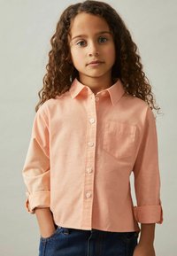 Light peach button-up shirt with a collar, long sleeves rolled at cuffs, front pocket, textured fabric, worn with dark denim jeans.
