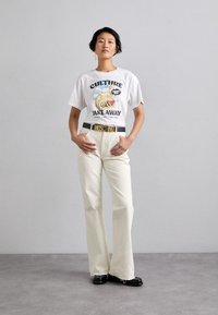 Balts T-krekls ar krāsainu grafisko tekstu "KULTŪRA ŅEM PAKAĻ", apvienots ar krēmkrāsas platām biksēm un melnu jostu, kurai ir "MOSCHINO" zelta burti.