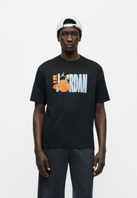 Μαύρο βαμβακερό μπλουζάκι με πολύχρωμο γραφικό "AIR JORDAN" και εικονογράφηση καλαθοσφαίρισης στο μπροστινό μέρος. Λαιμόκοψη τύπου crew και κοντά μανίκια.