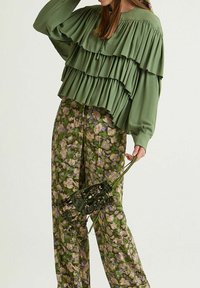 Blusa verde a strati con volants, abbinata a pantaloni stampati a fiori in verdi e viola. Accessori comprendono una piccola borsa decorata.