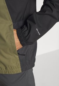 Chaqueta negra y verde oliva con puños elásticos y dobladillo ajustable. Presenta la marca "DRYVENT" en la manga; tela lisa y resistente al agua.