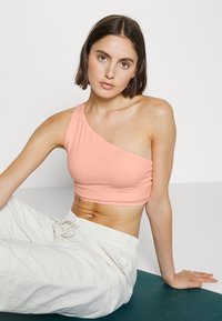 Top cropped assimétrico em pêssego claro com uma única alça no ombro, apresentando um tecido suave e textura franzida, combinado com calças brancas de corte descontraído.