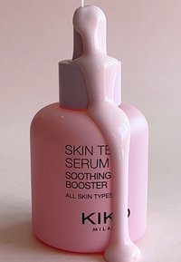 Rosa serumflaska med glansig yta, utrustad med en slät, förlängd pumpkork och svart textetikett; vätskan droppar ned längs sidan.