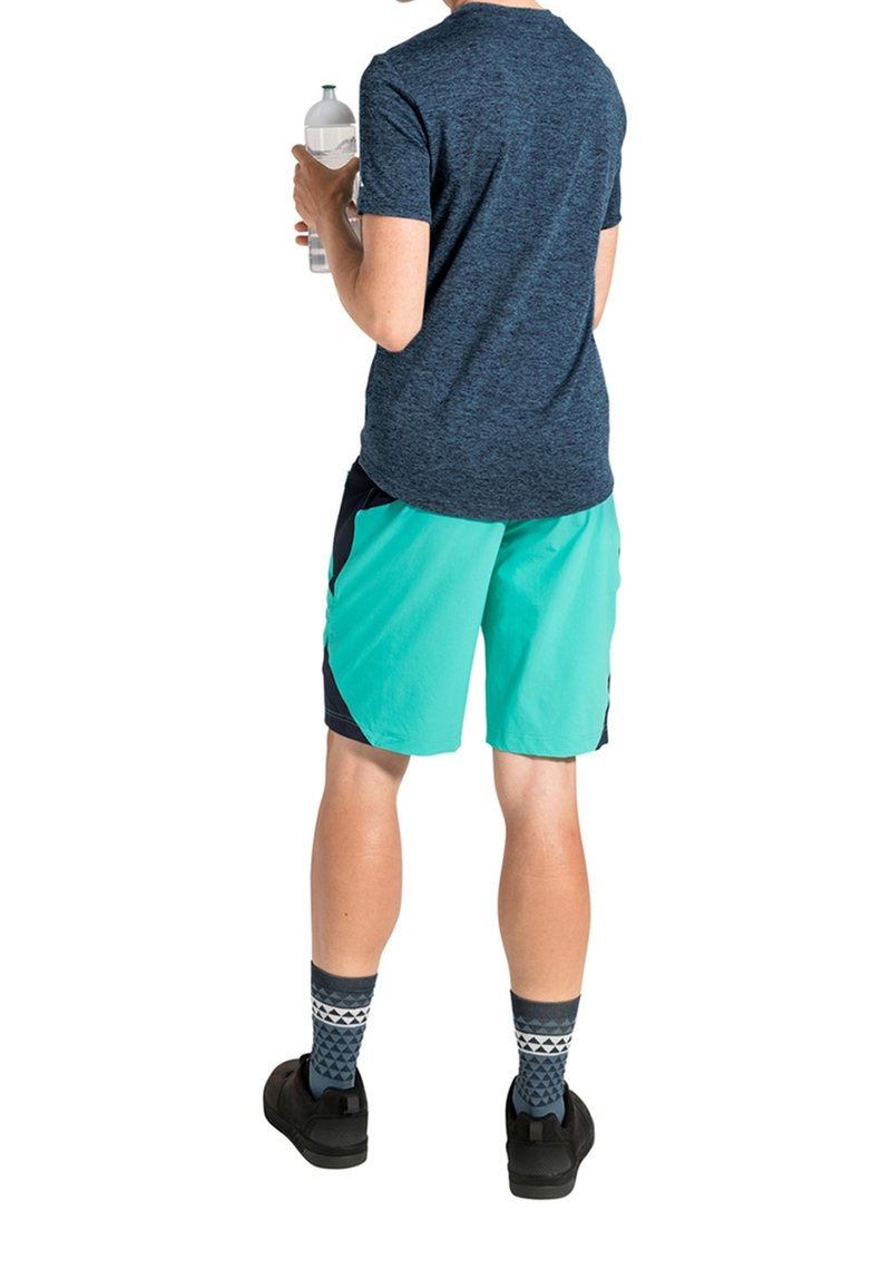 Personne portant un t-shirt bleu foncé et un short turquoise tient une bouteille d'eau transparente, debout dos au spectateur sur fond blanc.