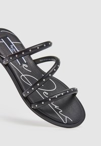 Sandale plate noire avec trois brides cloutées et logo blanc en écriture cursive sur la semelle, présentée sur un fond uni.