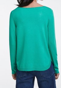 Femme portant un pull à manches longues vert vif et un jean bleu foncé, vue de dos sur fond blanc.