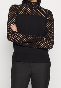 Haut en maille noire avec un col montant et des manches longues, présentant des ouvertures sur toute la partie supérieure et un tissu uni au niveau de l'ourlet inférieur.