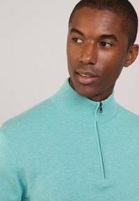Pullover lavorato a maglia di color azzurro chiaro con colletto alto zip. Tessuto a maglia fitta, polsini a costine e corpo liscio.