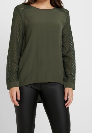 Femme portant un haut à manches longues vert olive avec des manches transparentes à motifs et un pantalon en simili-cuir noir, sur un fond uni.