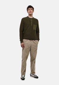 Pull en vert foncé texturé avec une petite poche, col à boutons ; associé à un pantalon en velours côtelé beige clair et des baskets noir et blanc.