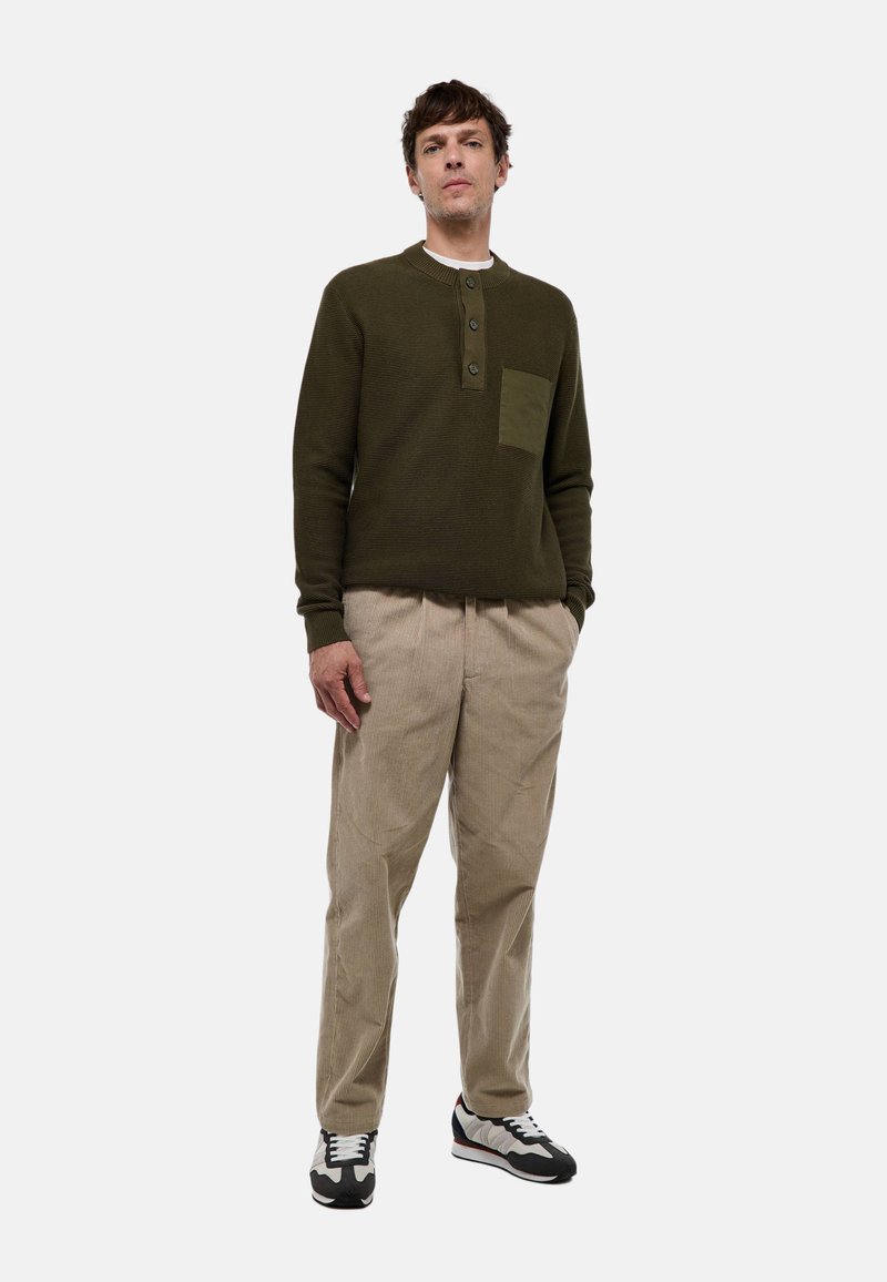 Pull en vert foncé texturé avec une petite poche, col à boutons ; associé à un pantalon en velours côtelé beige clair et des baskets noir et blanc.