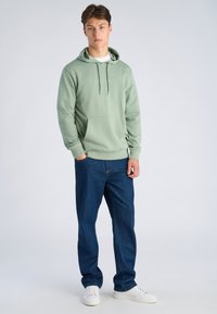 Jonge man die staat met handen in zijn zakken, draagt een lichtgroene hoodie, donkerblauwe spijkerbroek en witte sneakers tegen een effen achtergrond.