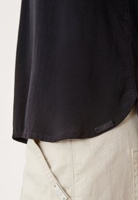 Maglietta nera a maniche corte con orlo arrotondato e un leggero spacco laterale, caratterizzata da un tessuto testurizzato e un piccolo etichetta del marchio in basso.