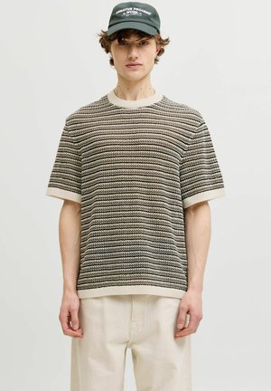 Jonge man met een zwarte pet en een beige, grijs en zwart gestreepte korte mouwen gebreide trui met ecru broek tegen een effen achtergrond.