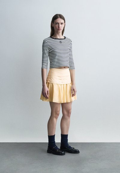 Νεαρή γυναίκα που στέκεται φορώντας μαύρο και λευκό ριγέ κοντό τοπ, ωχρή κίτρινη φούστα, μαύρες κάλτσες και μαύρα μοκασίνια μπροστά σε έναν απλό τοίχο.