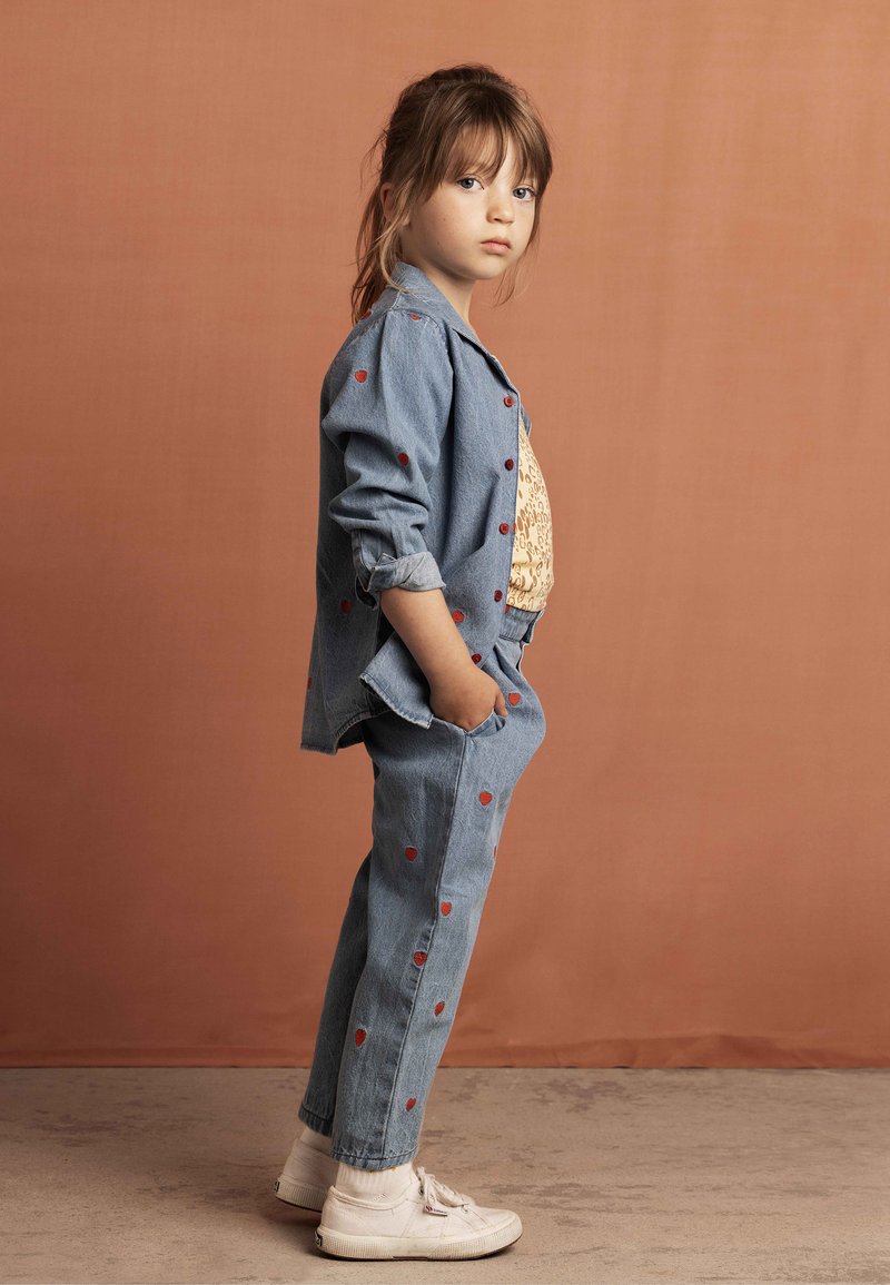 Denim outfit featuring a light blue jacket and pants with red heart patterns. Paired with white sneakers. Set against an orange background.