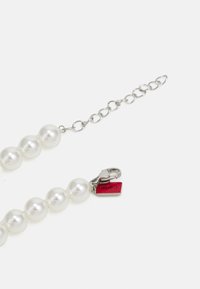 Witte parelketting met een zilveren ketting, bestaande uit ronde parels en een rood label met "HUGO" naast een zilveren kreeftsluiting.