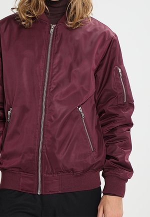 Personne portant une veste bomber bordeaux avec des poches zippées sur le devant et les manches, debout devant un fond clair uni.